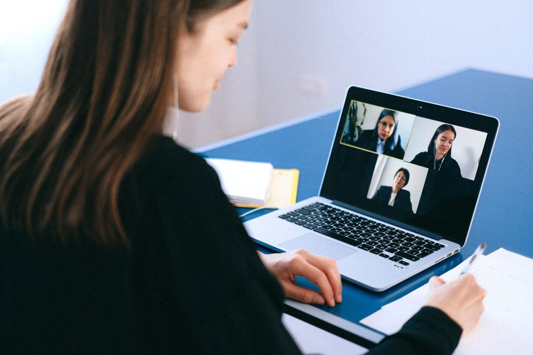 A person on a video call at a laptop in a home office, headset on and notes beside the keyboard